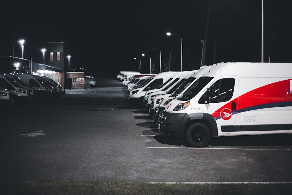 Nighttime view of a parking lot with a row of white delivery vans and several passenger cars parked on the left side. The vans are aligned in a line, with the closest one displaying a red and blue logo, and are situated in designated parking spaces on an asphalt surface. Bright streetlights illuminate the area, casting light over the vehicles and pavement. The background features a building with lit windows and additional parked vehicles, suggesting a commercial or courier depot environment. During home relocation or furniture transport, such parking arrangements are often used for loading or unloading belongings. Man with Van Petts Wood regularly supports moving logistics, including parking management and transportation, to ensure compliance with local parking rules for moving vans, particularly during busy moving days.