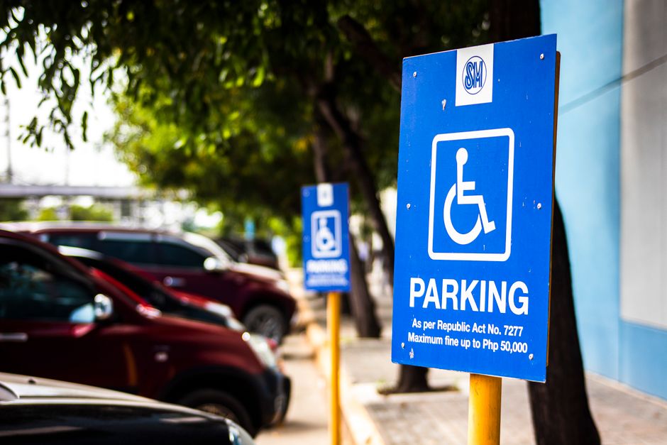 A close-up view of a bright blue parking sign indicating a designated space for wheelchair users, with white text and symbols. The sign is mounted on a yellow metal pole and features the international accessibility symbol. In the background, there are several parked cars, primarily in shades of black and red, situated along a paved street. The environment is outdoors with trees providing some greenery and shade, and a building with a grey wall partially visible on the right. The lighting is natural daylight, highlighting the sign and surrounding vehicles. Occasionally, moving vans or service vehicles related to house removals might park nearby during moving logistics, which Man with Van Petts Wood facilitates through proper parking in accordance with local regulations and parking rules for home relocations, evident from the context of parking signs for transport and moving services.