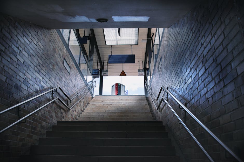 An upward view of a staircase inside a modern station, with dark tiled steps flanked by metal handrails on both sides, leading to an open exit area illuminated by natural daylight. The walls are const