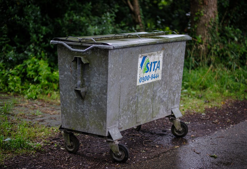 A large, rectangular metal waste container with four black wheels positioned on a paved driveway, partially on a patch of dirt and grass. The container has a hinged lid on top and a white sticker with blue and green writing, indicating the waste collection company 'SITA' along with a contact number. The background consists of green foliage and trees lining the edge of the property. The scene appears to be outdoors during daytime, with natural lighting highlighting the weathered surface of the container. The image is related to house removals and moving logistics, illustrating the type of external waste disposal equipment used during home relocations managed by Man with Van Petts Wood, supporting the understanding of proper pavement and skip rules in Petts Wood.