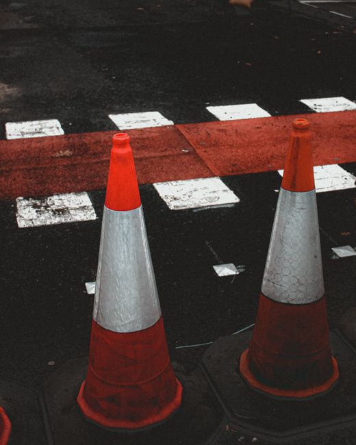 Two orange and white traffic cones positioned on a pavement next to a red and white striped pedestrian crossing area. The cones are placed near the edge of the road, which has a section of tarmac and painted white dashed lines to mark the crossing. The cones likely serve to restrict access or signal a loading zone during home relocation or furniture transport activities. The background shows part of the dark road surface and faded road markings, indicating an outdoor street environment used for loading and unloading items during a house removal. This scene illustrates typical pavement and vehicle control measures associated with professional removals, provided by companies such as Man with Van Petts Wood, to facilitate safe and efficient moving procedures.
