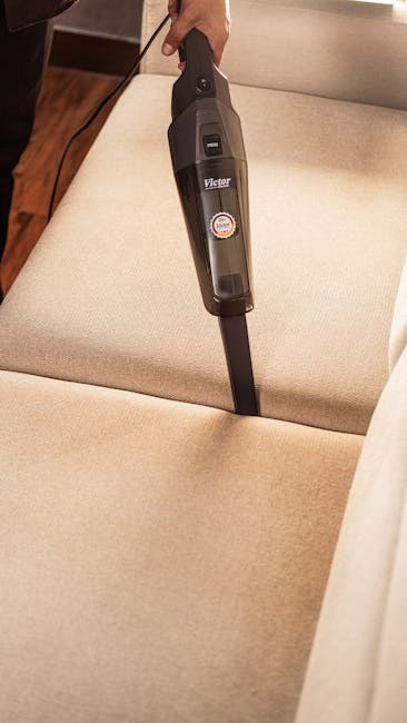 A person using a handheld vacuum cleaner to clean a beige fabric sofa, with the vacuum positioned centrally on the sofa's seat cushion. The vacuum has a black and grey body with a small red and white circular sticker, and a long, slim nozzle attachment. The sofa is situated inside a room with wooden flooring partially visible along the edges of the sofa, and the background shows a light-colored wall. The scene depicts a home cleaning or preparation process related to house removals or packing for a move, and the image is associated with professional removal services such as those offered by Man with Van Petts Wood, specializing in furniture transport and home relocation tasks.