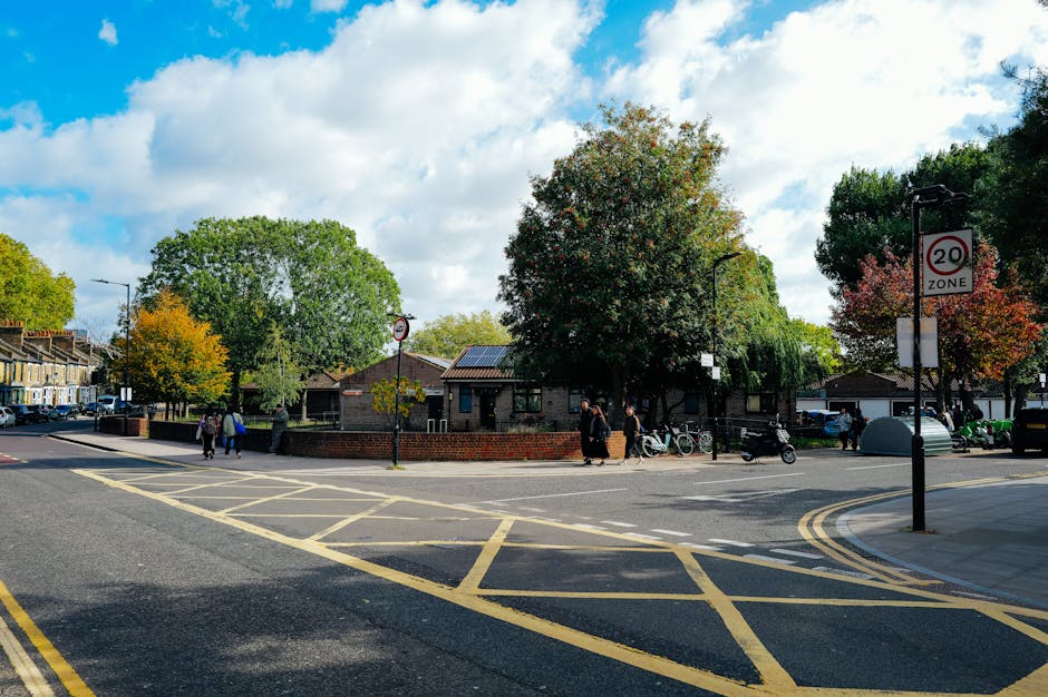 A street scene in Petts Wood showing a small public space with a brick wall surrounding a garden area containing large trees with green and autumnal foliage. Several pedestrians are walking along the pavement and crossing the road, some with bicycles parked nearby. Vehicles are parked along the curb, and a white van, likely used for home relocation or furniture transport, is visible at the edge of the scene. The sky is partly cloudy with patches of blue, and there are lampposts along the street, including one with a 20 mph zone sign. This setting illustrates an urban environment suitable for house removals and moving services offered by Man with Van Petts Wood, with a focus on local area logistics and community surroundings.