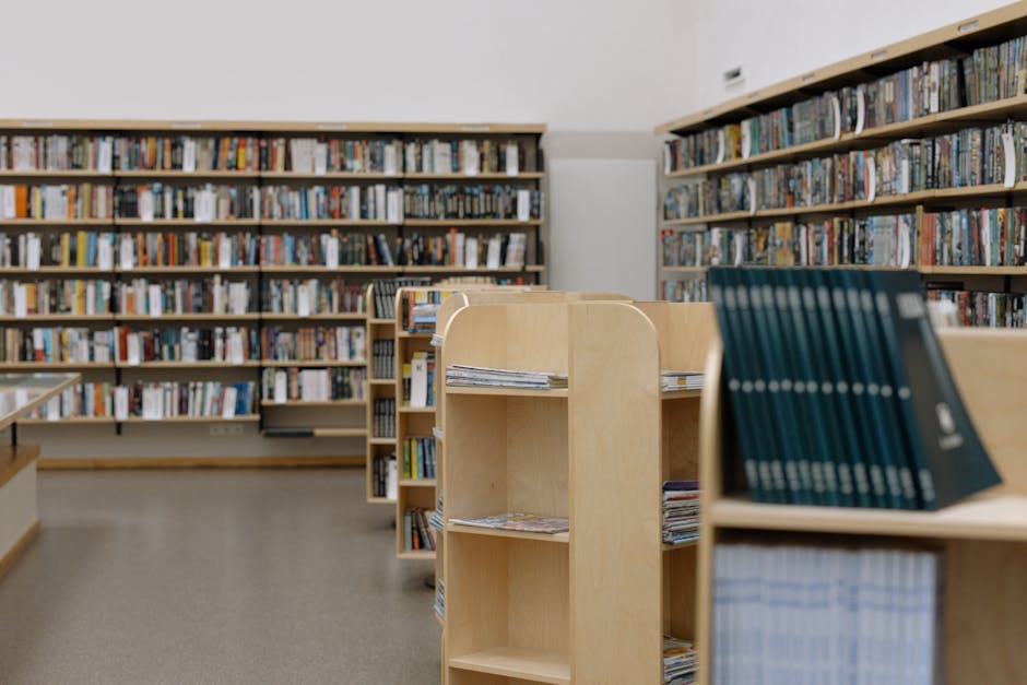 The image shows a long aisle within a library, featuring tall wooden bookshelves filled with neatly arranged books, mostly paperbacks and hardcover editions with visible labels on the spine. The wooden shelves are a warm tone, contrasting with the dark hardwood floor that runs the length of the corridor. The shelves are divided into sections, some with small digital or printed labels indicating categories. Overhead, soft lighting illuminates the space, creating a calm and organized atmosphere typical of library environments. In the background, a person is visible actively transporting or organizing books, possibly involved in home or institutional library relocation or packing processes. The scene captures the interior of a library or shared space used for reading and study, during a moving or reorganization process that might be supported by companies like Man with Van Petts Wood, who specialise in removals and furniture transport. The setting emphasizes careful handling of library items, with no visible packing materials, but the context suggests ongoing packing or moving logistics.