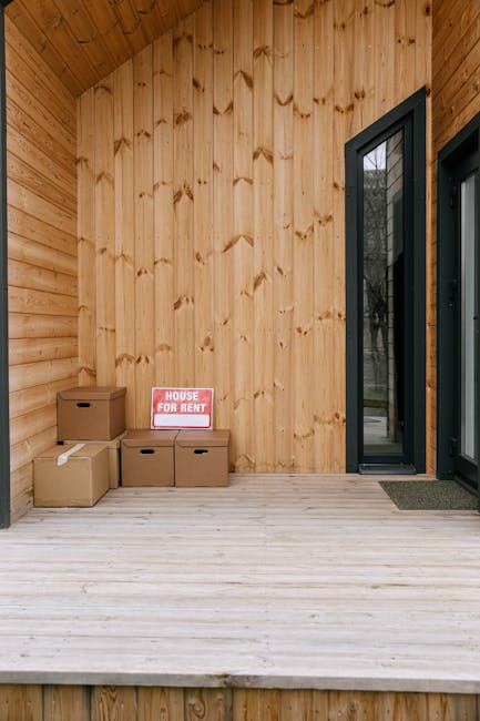 A man and a woman, both dressed in casual clothing, are carrying cardboard boxes towards a modern wooden house with large glass sliding doors, outside a property during daytime. The man is wearing a b