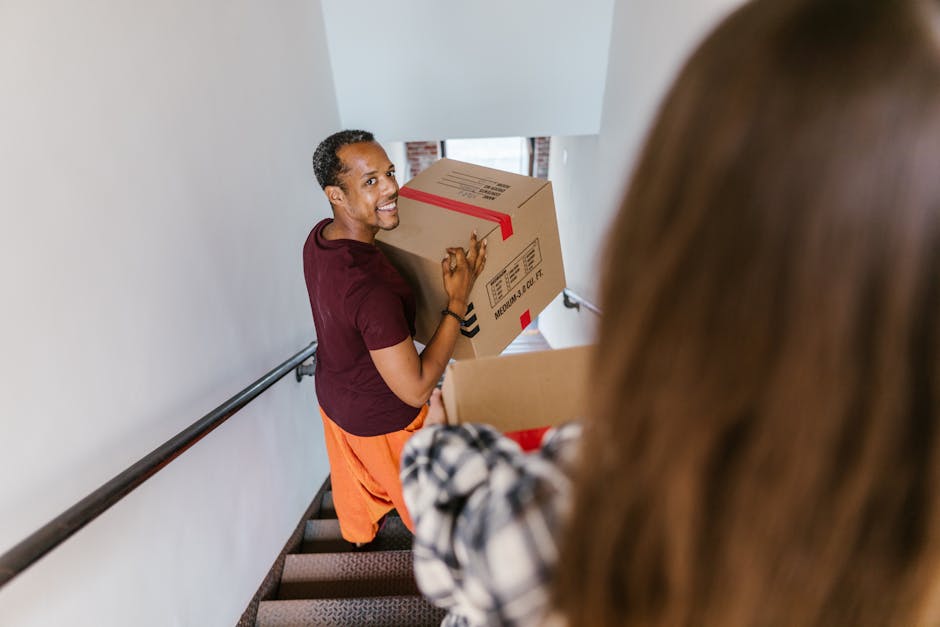 A man with dark skin and short curly hair, wearing a maroon t-shirt and orange pants, is seen smiling while carrying a large cardboard box with red tape and printed labels. He is ascending a staircase inside a house during a home relocation process, with a handrail on his right side and a narrow corridor wall on his left. In the foreground, a woman with long brown hair, dressed in a black and white checkered shirt, is holding another cardboard box with red tape, assisting in the packing and moving activity. The interior space has white walls, and a window with natural light illuminates the area, indicating daytime. The scene captures a functional loading process typical of furniture transport and packing during a house removal, reflecting the logistics involved in professional moving services like those provided by Man with Van Petts Wood, with attention to safe handling of packed items within a home environment.