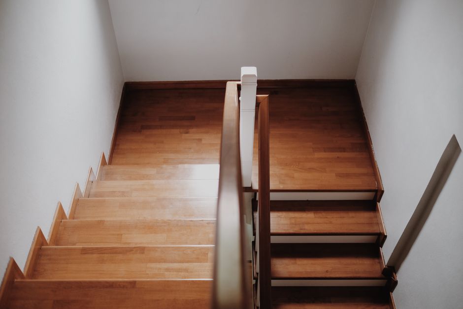 A top-down view of a wooden staircase inside a residential property, showing two sections separated by a landing; the lower section has polished wooden steps with a white safety railing on the left side, and the upper section continues with similar wooden steps accompanied by a wooden handrail on the right. The staircase is enclosed by white-painted walls, and natural light illuminates the space, highlighting the smooth surface of the wooden stairs. This indoor setting reflects the environment where house removals and furniture transport processes take place, often involving careful handling and packing of items such as boxes and furniture components, which are not visible in this specific image. The image exemplifies the typical internal environment encountered during home relocation projects managed by companies like Man with Van Petts Wood.