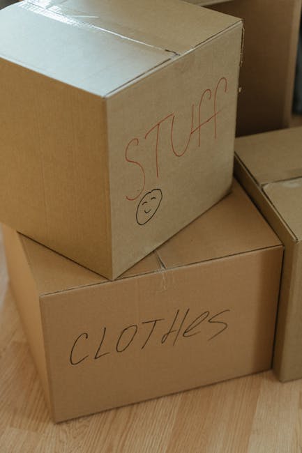 A close-up view of two medium-sized cardboard boxes stacked on a wooden floor, prepared for home relocation. The top box has the word 'STUFF' written in red marker, along with a simple smiley face underneath and a small circle. The lower box is labeled 'CLOTHES' in black marker. Both boxes are sealed with packing tape and are situated next to other partially visible boxes, indicating an organized packing process. The background shows additional boxes and a glimpse of a doorway, suggesting the boxes are inside a room ready for transportation or loading onto a moving vehicle, which is part of the furniture transport and packing services offered by Man with Van Petts Wood as they assist with home removals.