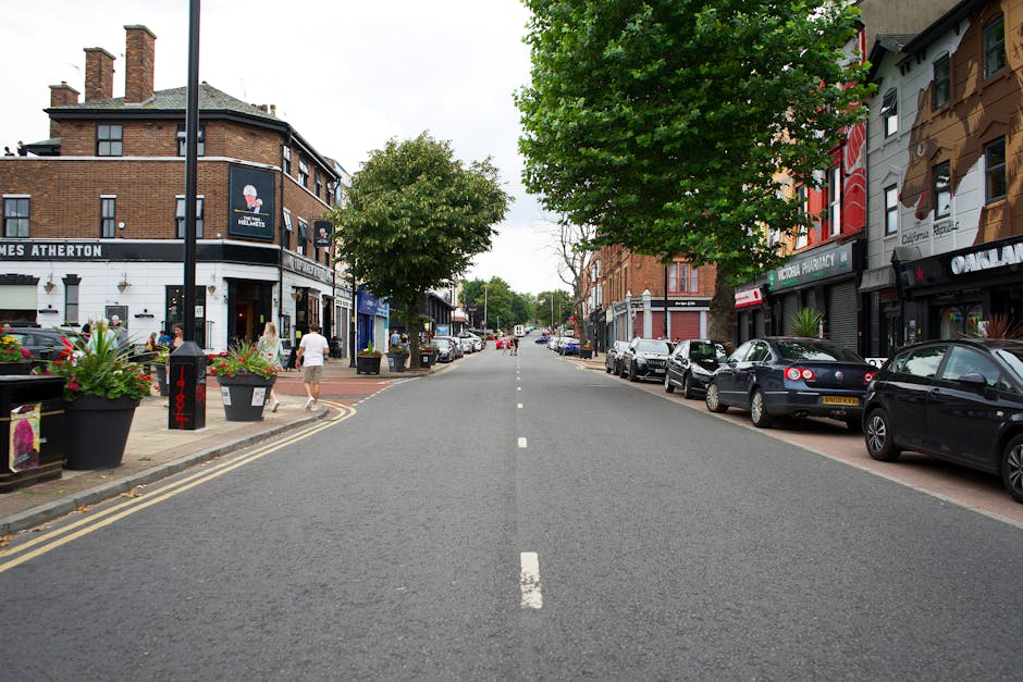 A quiet, urban street scene during daytime, with a clear view down the road lined with parked cars on both sides. On the left side, there are large planters with pink flowers and a black lamppost, with pedestrians walking along the sidewalk, some carrying bags or engaged in conversation. The buildings feature a mix of brick and painted facades, including shops and cafes, with visible signage such as 'James Atherton' and other storefronts. On the right side, more parked vehicles are visible, with a row of modern cars in various colors. Several trees with lush green foliage line the street, providing shade and a natural element to the scene. The road itself is paved with asphalt, marked with a central dashed white line, and appears to be in a busy part of town suitable for home relocation or furniture transport activities, consistent with the services offered by Man with Van Petts Wood. The overall environment is well-lit with natural daylight, emphasizing the urban setting for loading and moving processes.
