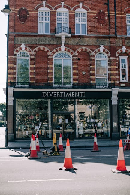 The image shows the façade of a multi-storey brick building with white window frames and decorative arch details above the windows. On the ground floor, there is a shop with large glass windows and a black awning displaying the name 'Divertimenti' in white letters. In front of the shop, the street is sectioned off with orange and white traffic cones, and there is a small area marked by yellow and black barriers, likely indicating a designated loading zone for home relocation or furniture transport. The scene is captured during daylight with natural lighting, and there are no people visible in the image. This setting illustrates an urban environment suitable for loading and unloading goods during a house removal or moving process, with support from professional services like Man with Van Petts Wood providing efficient packing and transportation solutions.